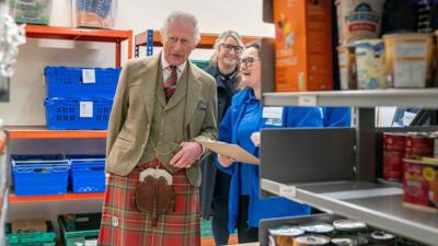 King Charles in a red kilt and beige jacket and waistcoat in a foodbank in Alloa. There are shelves with cans of food on them in the foreground.