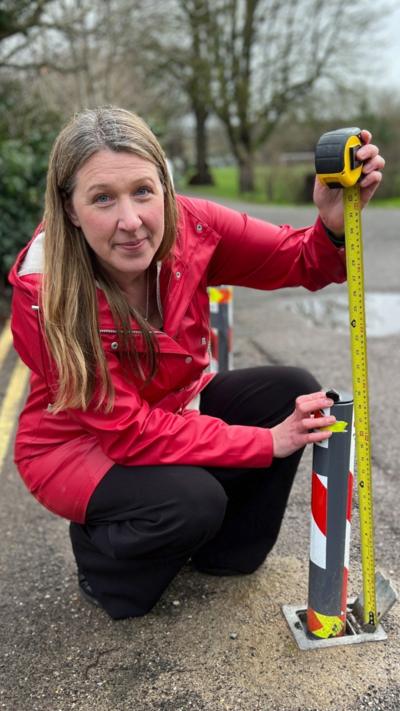 A woman is wearing a red coat, kneeling down to compare the height of a short bollard with a measuring tape.