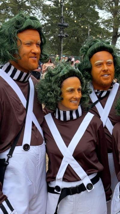 Kendal Calling festival goer dressed in a green wigs and orange face makeup based on Charlie and the Chocolate Factory. Three people are visible wearing brown costumes with white stripes. 