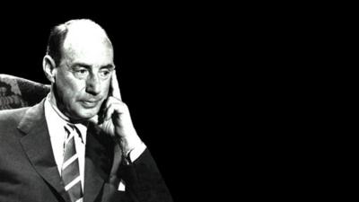 Black and white image of John Crawley sitting in chair on left of image.  he is holding the side of his face with his hand.  Wearing a tie and suit