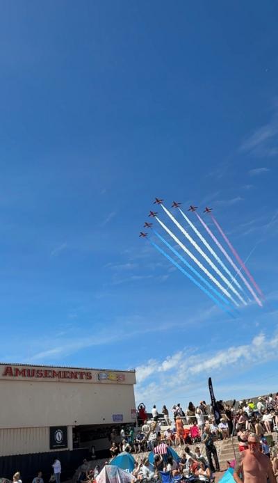 Seven Red Arrows planes fly in formation over Paignton seafront in a blue sky with a few white fluffy clouds. There is a crowd of people gathered on the beach looking up at the sky