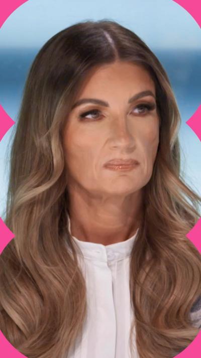 Karren Brady has long brown wavy hair and wears a white shirt. She looks stern.