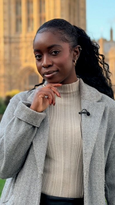 Jonelle Awomoyi looks at the camera resting her chin on her hand in front of Houses of Parliament. She wears a slick ponytail, a beige jumper and a grey coat.