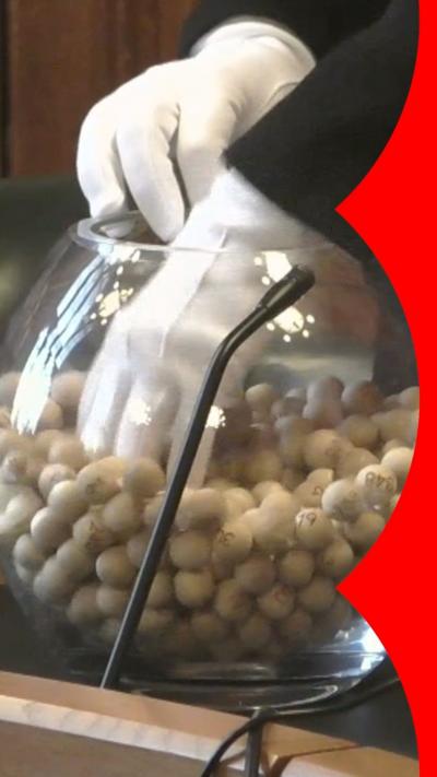 A white gloved-hand reaching into in a glass bowl full of small, numbered balls