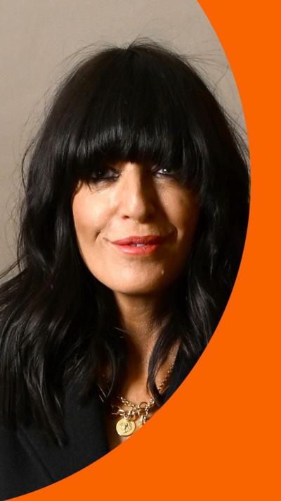 Claudia Winkleman wearing black clothes and gold necklace looking at camera.
