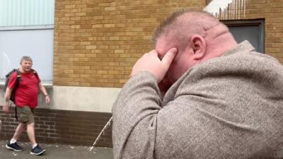 Side profile of Bernard McDonagh in a street. He holds his hand to his face covering his eyes. He has fine short hair and wears a brown tweed suit jacket. A man can be seen in the background wearing a red t shirt, cargo shorts and black trainers. 