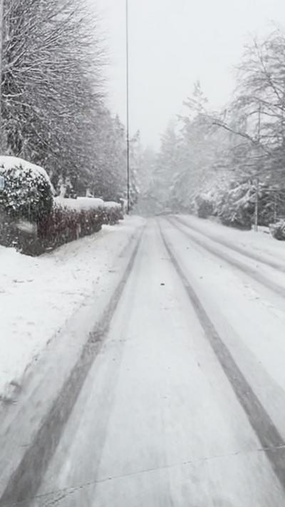 A snowy image taken from the front window of a car. The road in front is covered in snow, as well as the pavements and the trees in the distance. There is a person walking in a white jacket and black trousers on the pavement on the left hand side of the image.