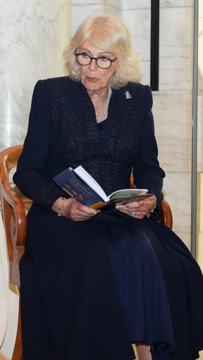 the Queen, wearing glasses, a navy blue gown and jacket, sits on a wooden chair holding a book. Her head is tilted as she faces children off camera towards her right.