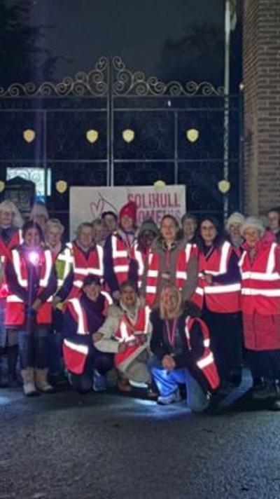 A large group of women together outside a park at night ahead of a walk. they are all wearing orange hi vis jackets