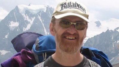 Marl wearing a white baseball cap that says 'Billabong', wearing sunglasses with a ginger beard. He's wearing a  backpack and hiking. There are snowy mountain peaks in the background.