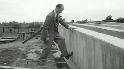 A man dressed in a suit, smoking a pipe, testing a large beam of concrete with his foot.