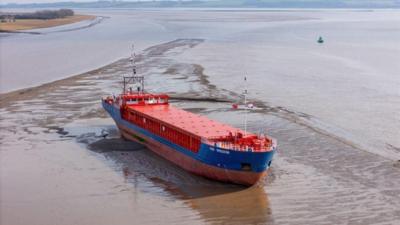 A red and blue cargo ship stuck in mud in the Humber, with brown waters to either side. The photo is taken from an aerial perspective.