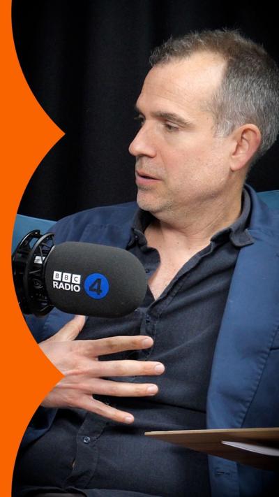 A man holds his hand over his chest. He is wearing a dark shirt with a blue jacket over it.