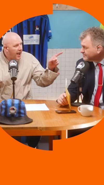 Two men sit at a table. The man on the left pointsat the other man angrily.