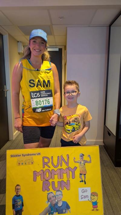A marathon runner in a yellow top with her young son who is also wearing yellow. They have a yellow poster which reads Run Mommy Run