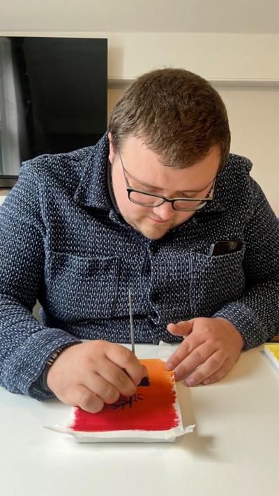 A man with brown hair and glasses sat at a table with a paintbrush. He is working on a painting that is red and orange. He's wearing a blue/grey patterned top