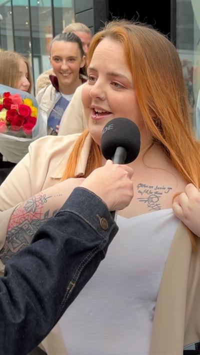 A woman pulls back her vest to reveal a tattoo on her upper chest