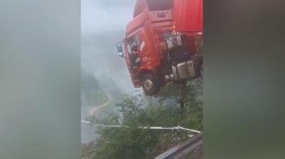 The cabin of a red lorry, with the driver leaning out of the open door, hangs over the edge of a broken bridge with a deep chasm below