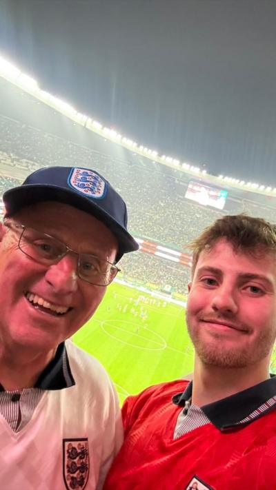 Jacob Allmendinger and his grandad, Geoff Golliker, wearing transparent rain ponchos hold a Hull City AFC St George’s flag in the stands of a large football stadium, overlooking the pitch before a match.