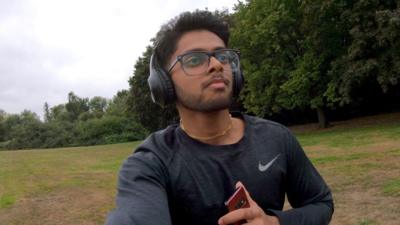 Mridul is running in a field, with his phone in hand and headphones on his head, while wearing glasses and a gold necklace