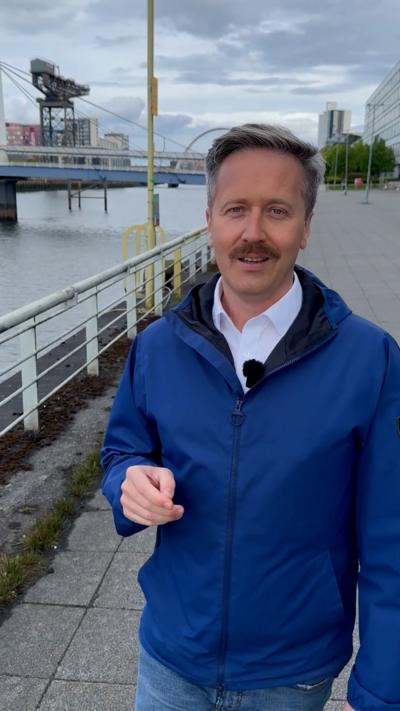 BBC Scotland weather presenter Christopher Blanchett, wearing blue coat and standing next to River Clyde