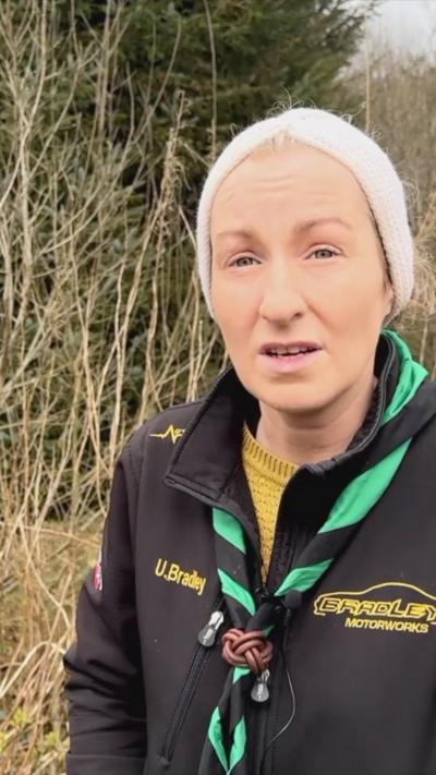 Ursula Bradley is wearing a knitted hat over the top of blonde hair, as well as a mustard-coloured knitted jumper with a black soft-shell top over it. Behind her foliage can be seen