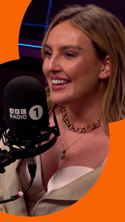 A woman, Perrie Edwards, smiling behind a Radio 1 mic. She wears a white crop top with beige jacket.