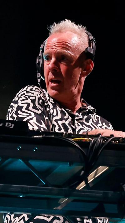 Fatboy Slim is on the decks. He has short grey hair which is longer on top. He has headphones on and is wearing a graphic black and white short.