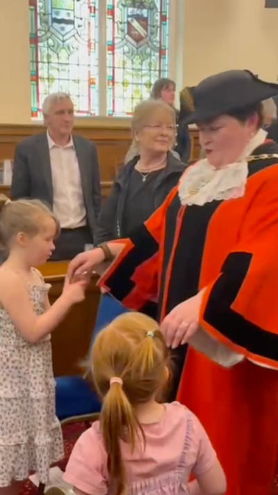 A person in a red ceremonial robe stands in a formal room with stained glass windows, interacting with two young children while several adults watch in the background.