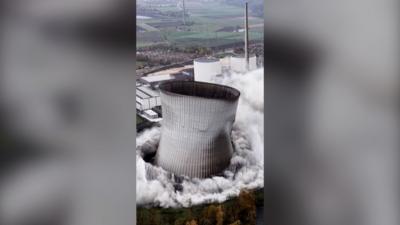 A grey concrete tower, mid-demolition, with plumes of white smoke at the bottom.