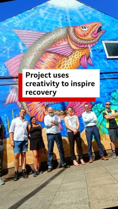 A colourful mural of a fish on the side of a building has a group of six people standing in front of it.