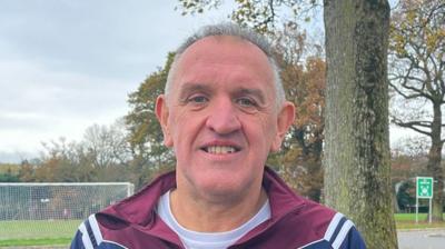 John Deery who has short grey/white hair and wearing a burgundy and navy tracksuit top with a white t-shirt underneath 