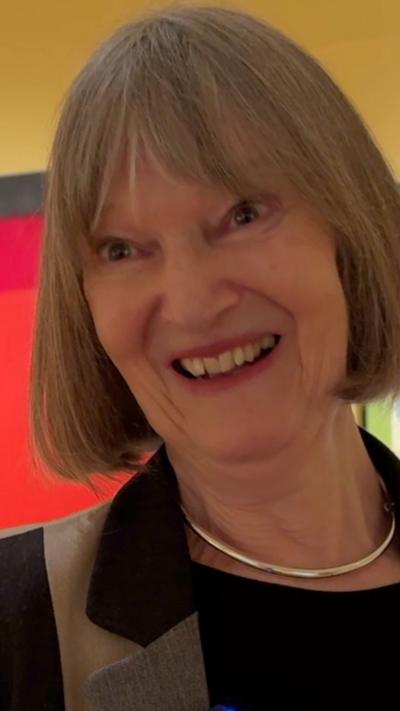 a smiling woman with her grey hair cut into a bob with a fringe. She is wearing a blazer 