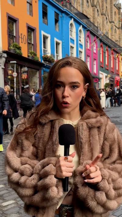 The video presenter faces the camera holding a black mic. She has dark brown hair and is wearing a light brown fur coat and cream top. Edinburgh's Victoria Street is in the background. Colourful shop fronts are visible.