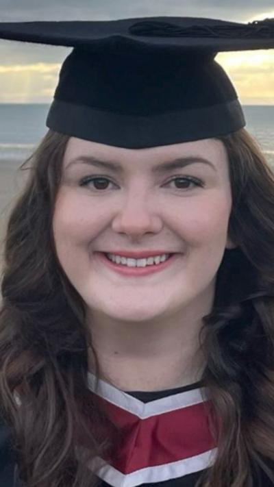 Caitlin with her graduation hat and gown on with the sea behind her. 