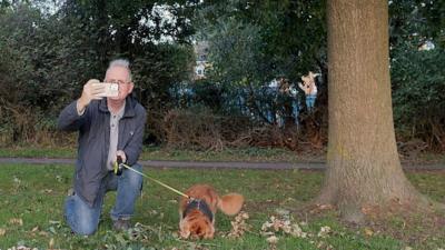 A man in a blue coat and blue jeans with a small brown dog next to a tree taking a photograph