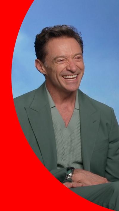 Hugh Jackman sits in front of a blue screen and smiles. He is wearing a green top and blazer. 