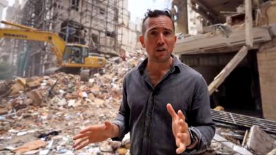 Middle East correspondent Hugo Bachega gestures while speaking to camera in front of a digger and rubble on a residential street in Beirut.
