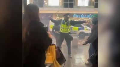 A police officer in a high-vis vest wields a baton at football fans running past shoppers on Glasgow's Argyle Street. A woman with long brown hair and shopping bags looks on. Football fans can be seen running past in the background.