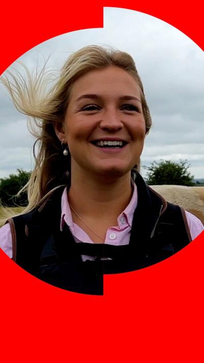 Farmer Harriet Cowan smiles while standing in a field - she has long blonde hair which is being blown by the wind