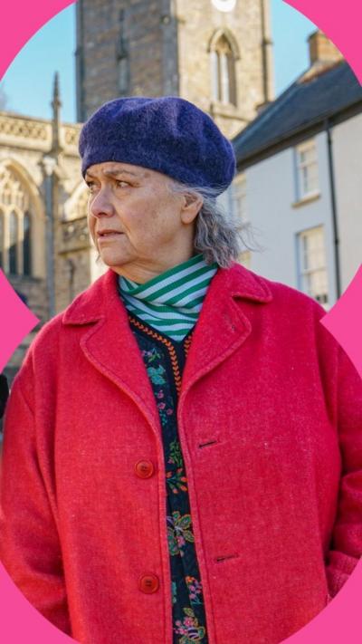 A woman wearing a red coat and purple beret looks to the side. A church is behind her 