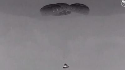 Black and white picture of capsule being hanging below three parachutes