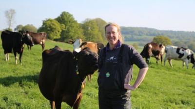 Dairy farmer Lorna Burdge stands in a field wearing overalls, with cows behind her