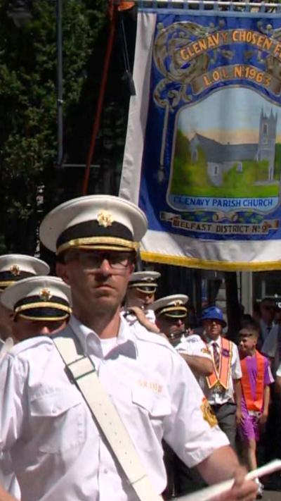 A band marches through Belfast, they are wearing white shirts and white caps. Behind them is a banner with a painting of a church on it