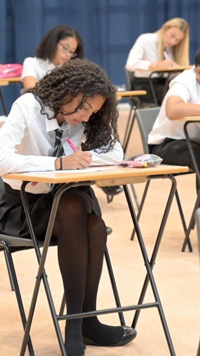 Teenagers sit at desks writing in an exam hall.