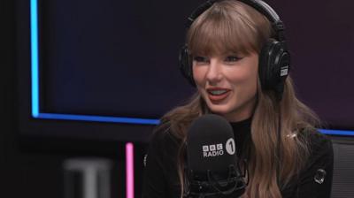 Taylor Swift wearing black headphones and in front of a BBC Radio 1 microphone