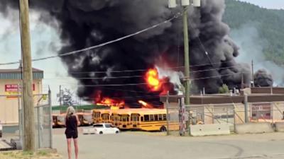 A woman stands in front of a raging fire and a huge plume of black smoke rising above yellow school buses