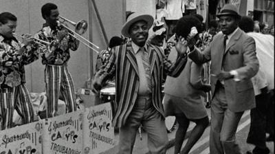 Black and white image of two men in suits dancing in the street to a calypso band.