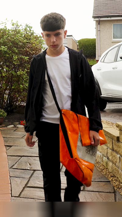 Boy with short brown hair wearing white T-shirt and dark tracksuit top, with orange satchel across his shoulder.