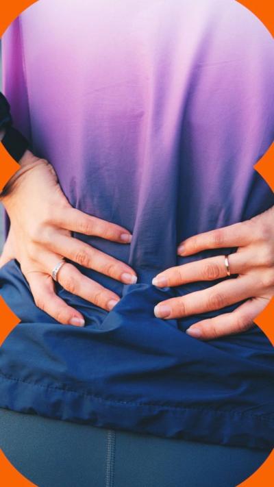 A woman holding her lower back, viewed from behind, wearing navy and purple jumper.